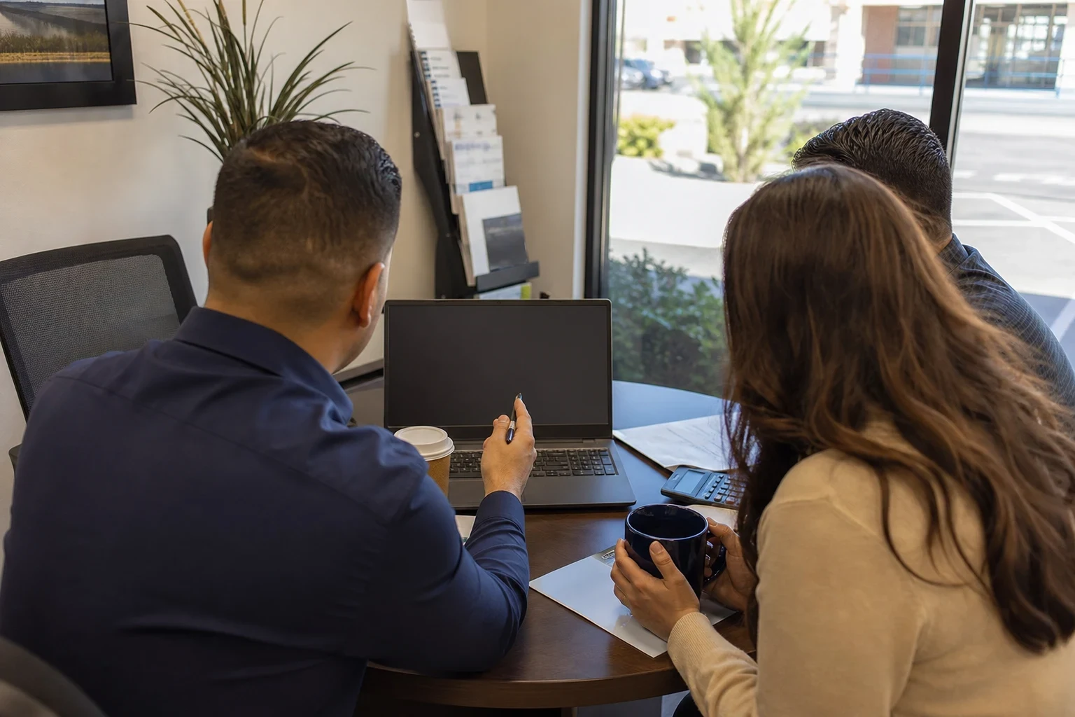 Agent in a navy shirt points a pen at a laptop while a couple sits with him at a small round office table, holding coffee, with a window to a parking lot behind
