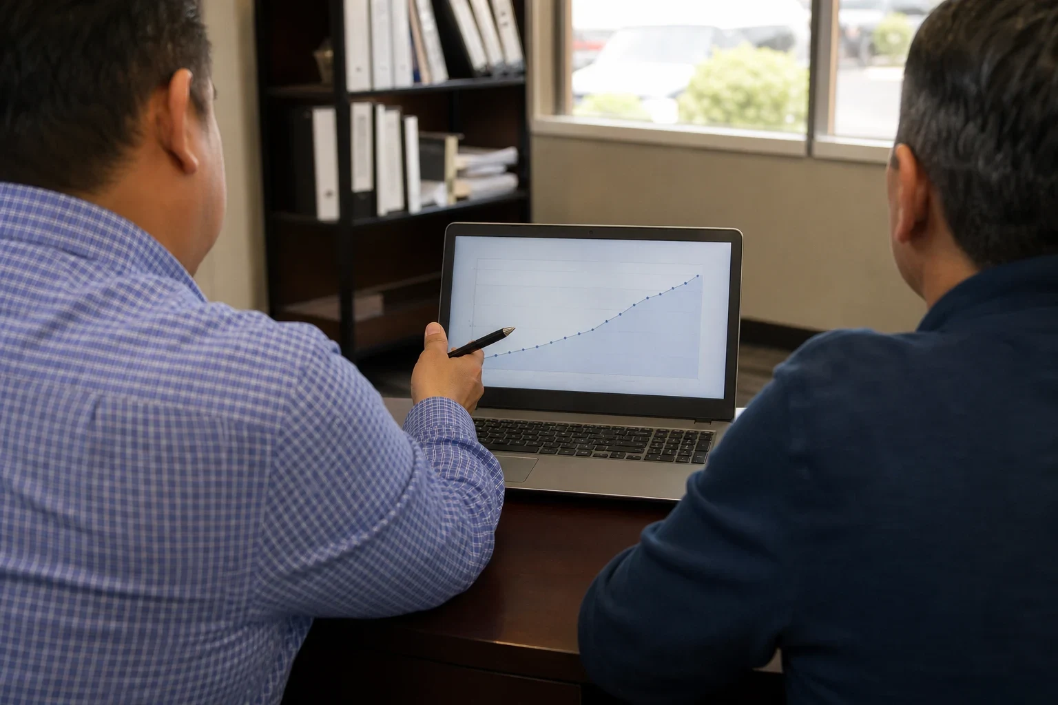 Two men seen from behind at an office desk, one pointing a pen at a laptop showing a steadily rising line chart, with a bookshelf of binders and a window behind